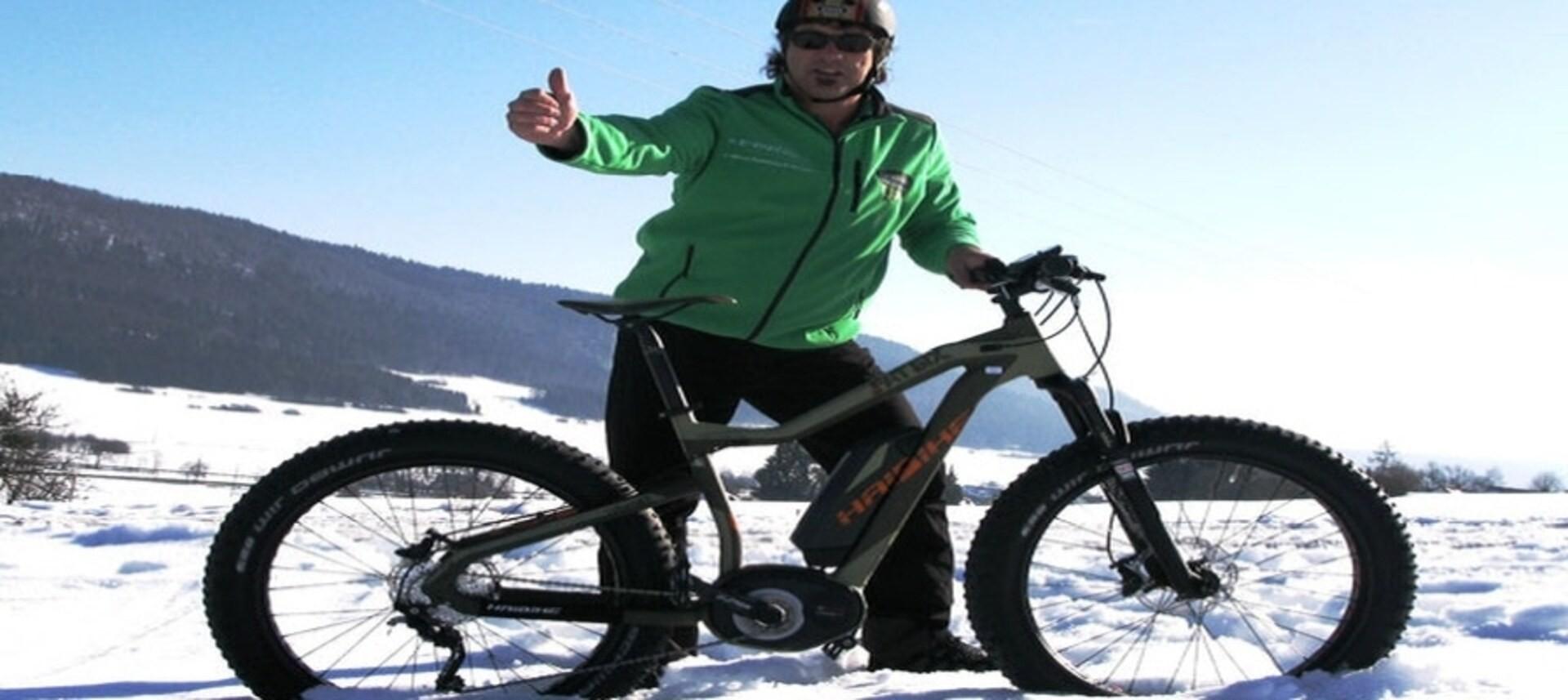 Fahrer auf Haibike XDURO Fatsix im Schnee, Daumen hoch, mit Helm und grüner Jacke, vor winterlicher Berglandschaft.