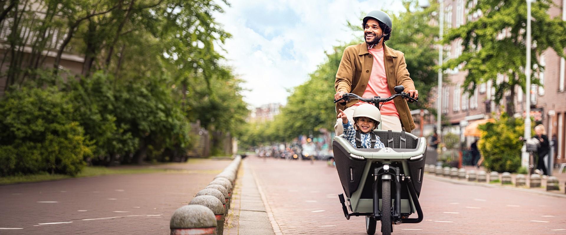 Ein Mann fährt mit einem Lastenrad durch die Stadt, vorne in der Transportbox sitzt ein Junge