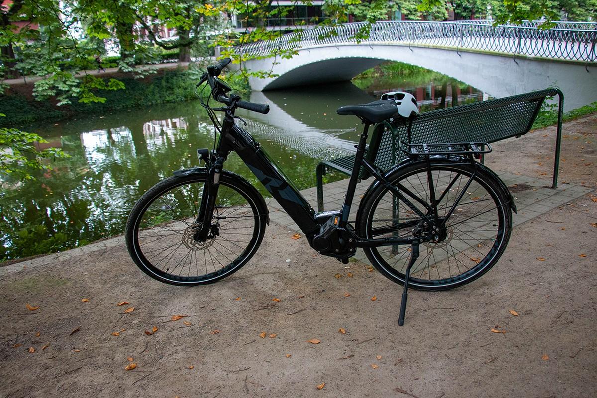 Das Bild zeigt ein Raymon Cityray e-Bike, das an einer Parkbank in der Nähe eines Gewässers abgestellt ist. Der Rah