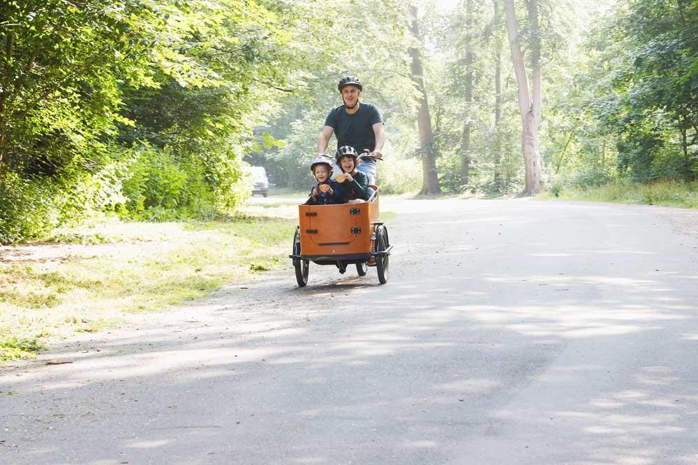 Ein Mann fährt mit zwei Kindern in der Transportbox des Babboe Lastenrads einen Waldweg entlang