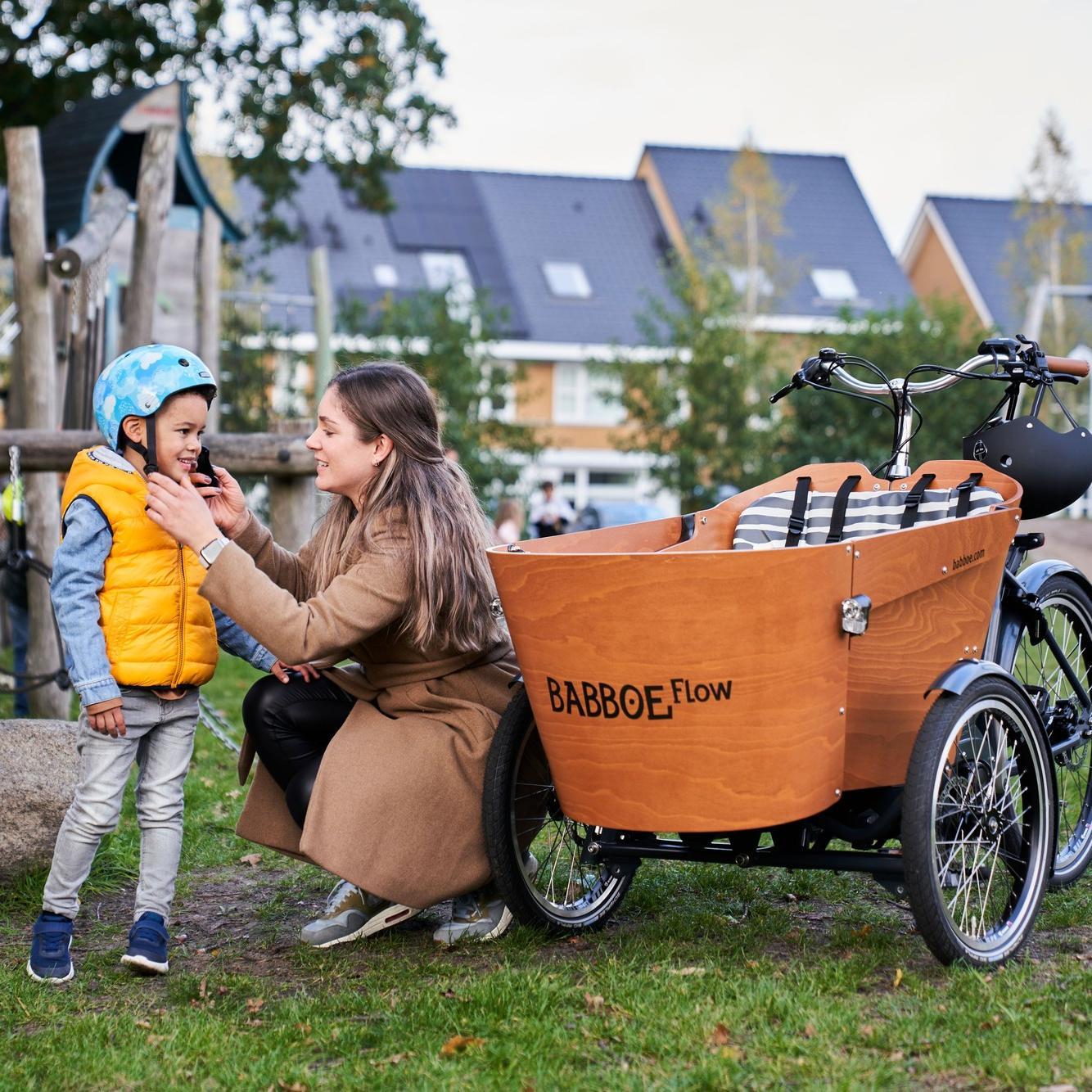 Eine Frau zieht einem Helm einem Kind an, daneben steht ein Babboe Flow Lastenrad