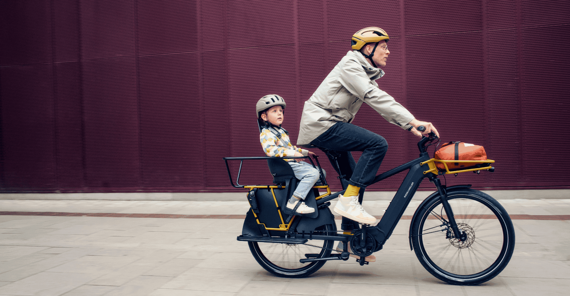 Ein Erwachsener fährt ein Lasten-E-Bike mit Kindersitz, in dem ein Kind mit Helm sitzt, vor roter Wand.
