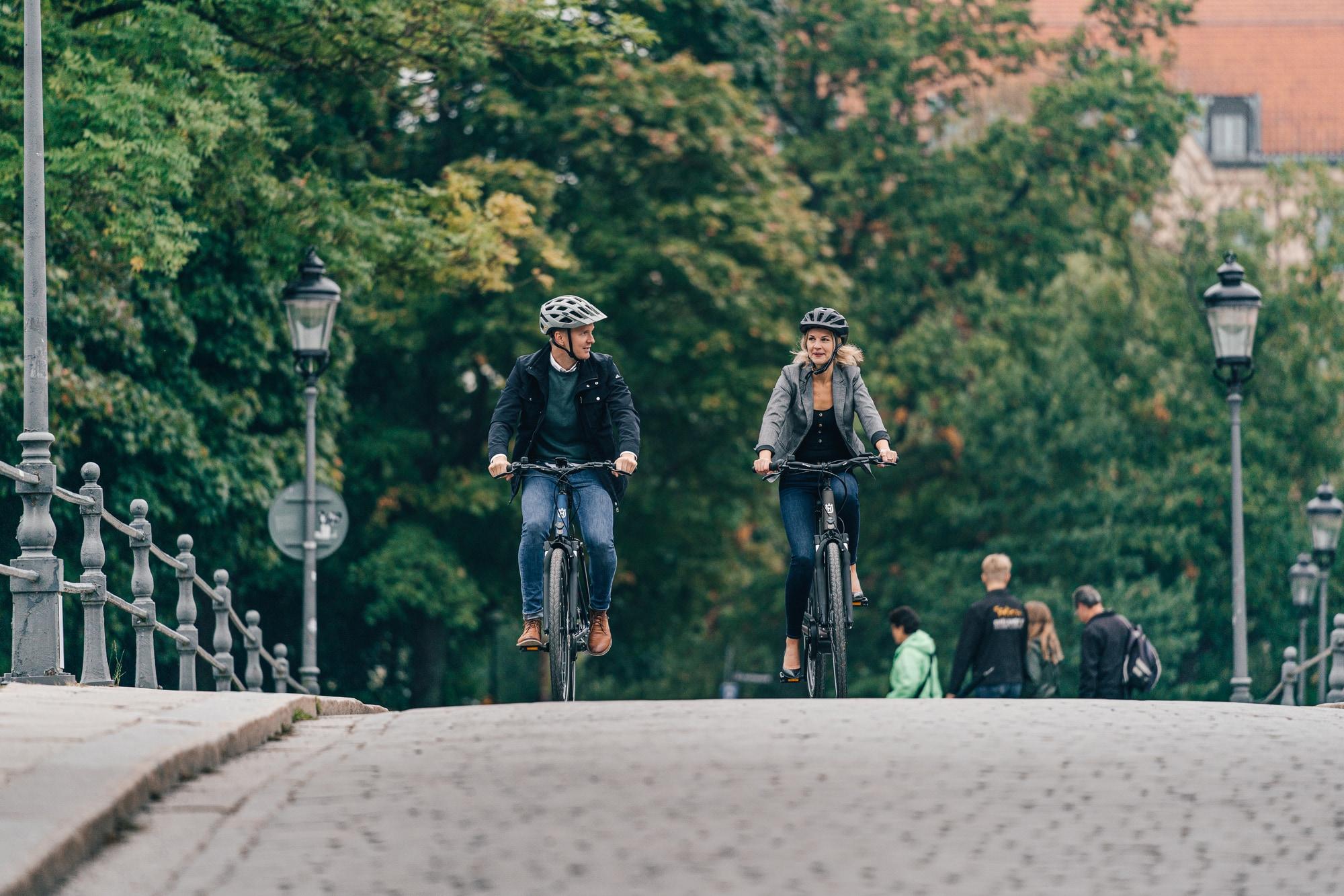 Zwei Personen fahren Eco City E-Bikes auf einer Brücke, umgeben von städtischem Grün.