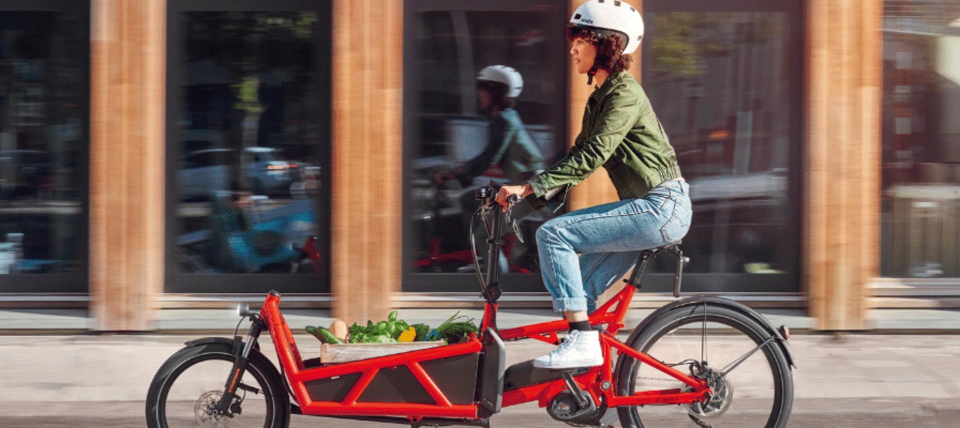 Das Bild zeigt eine Frau, die auf einem roten Lastenrad fährt.