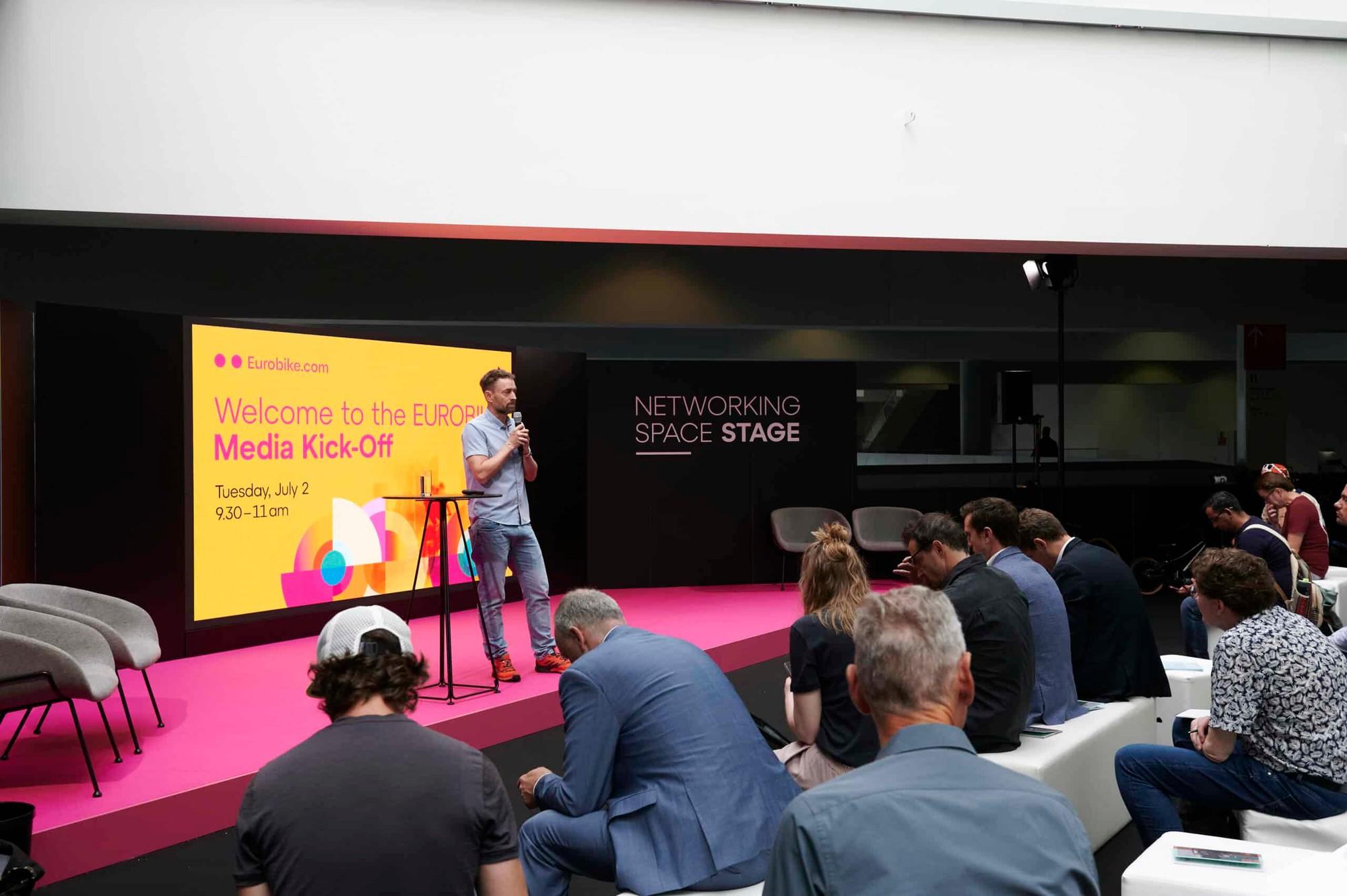 Präsentation auf pinkfarbener Bühne mit Redner, großem Bildschirm und aufmerksamem Publikum.