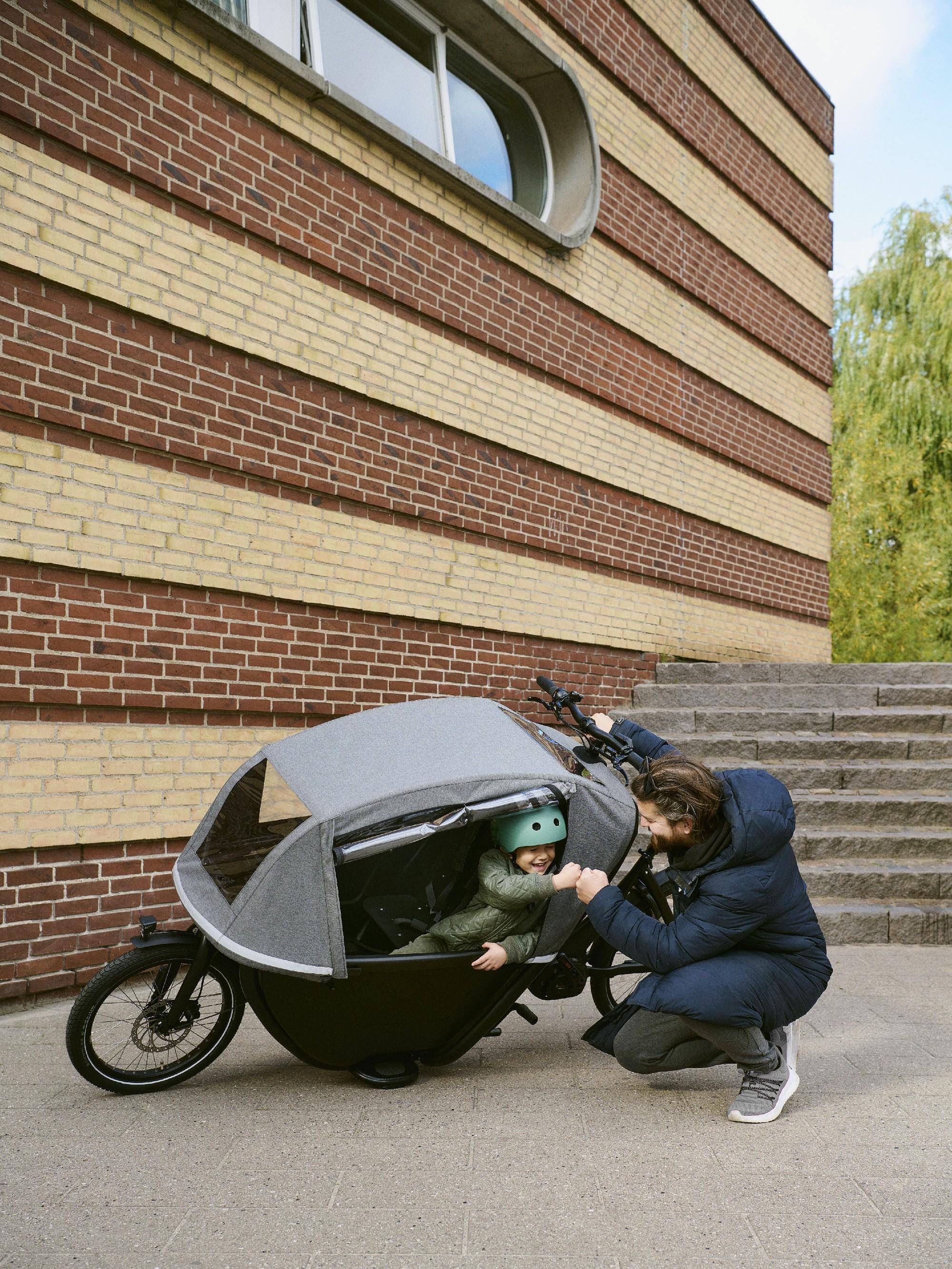 Ein Mann hockt neben einem Winther Lastenrad und schlägt bei einem Kind ein, welches in der Transportbox sitzt