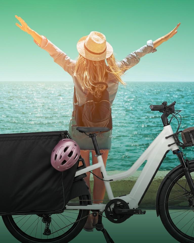 Eine Frau steht vor dem Wasser und breitet die Arme aus, hinter ihr ein e-Bike