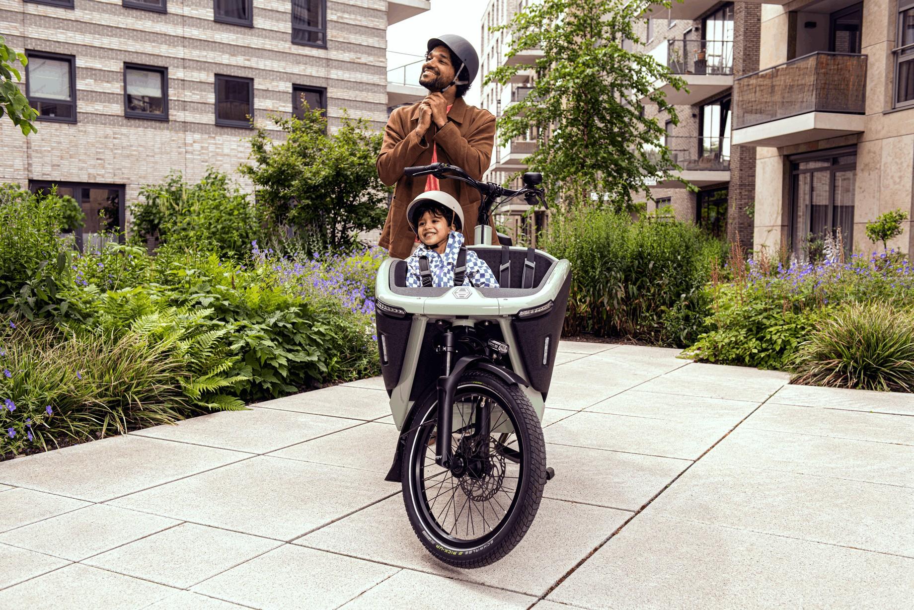 Vater mit Kind in einem Lastenrad auf einem Weg zwischen modernen Wohngebäuden.
