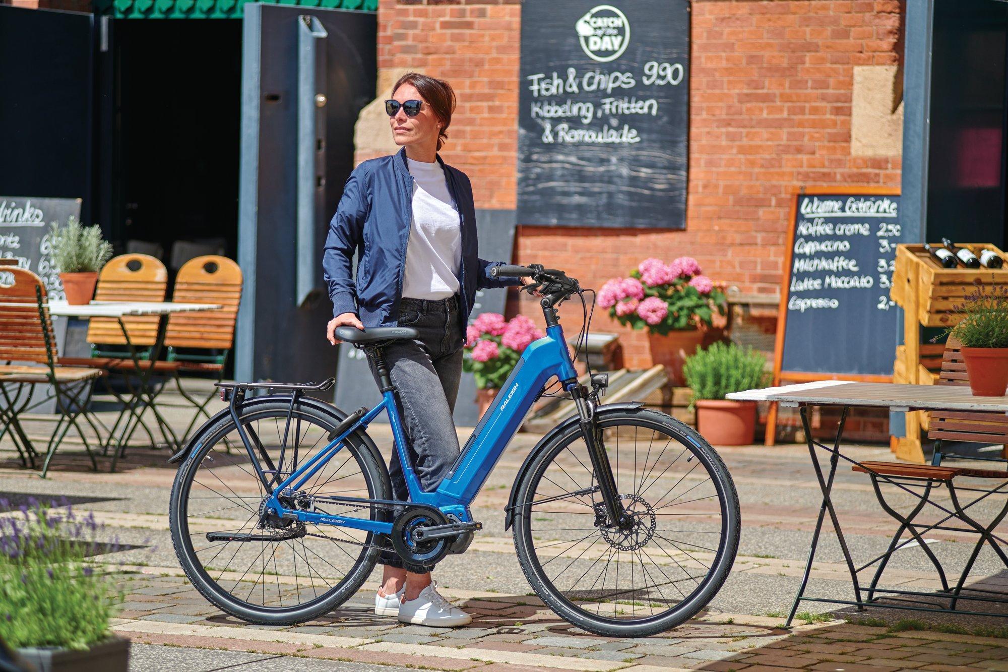 Eine Frau steht neben einem blauen E-Bike, das auf einem gepflasterten Platz vor einem Café parkt. Sie trägt Sonnen