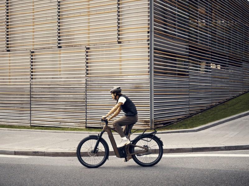 Ein Mann fährt auf dem Riese & Müller Delite5 GT Pinion HS e-Bike an einem modernen Gebäude eine Straße entlang
