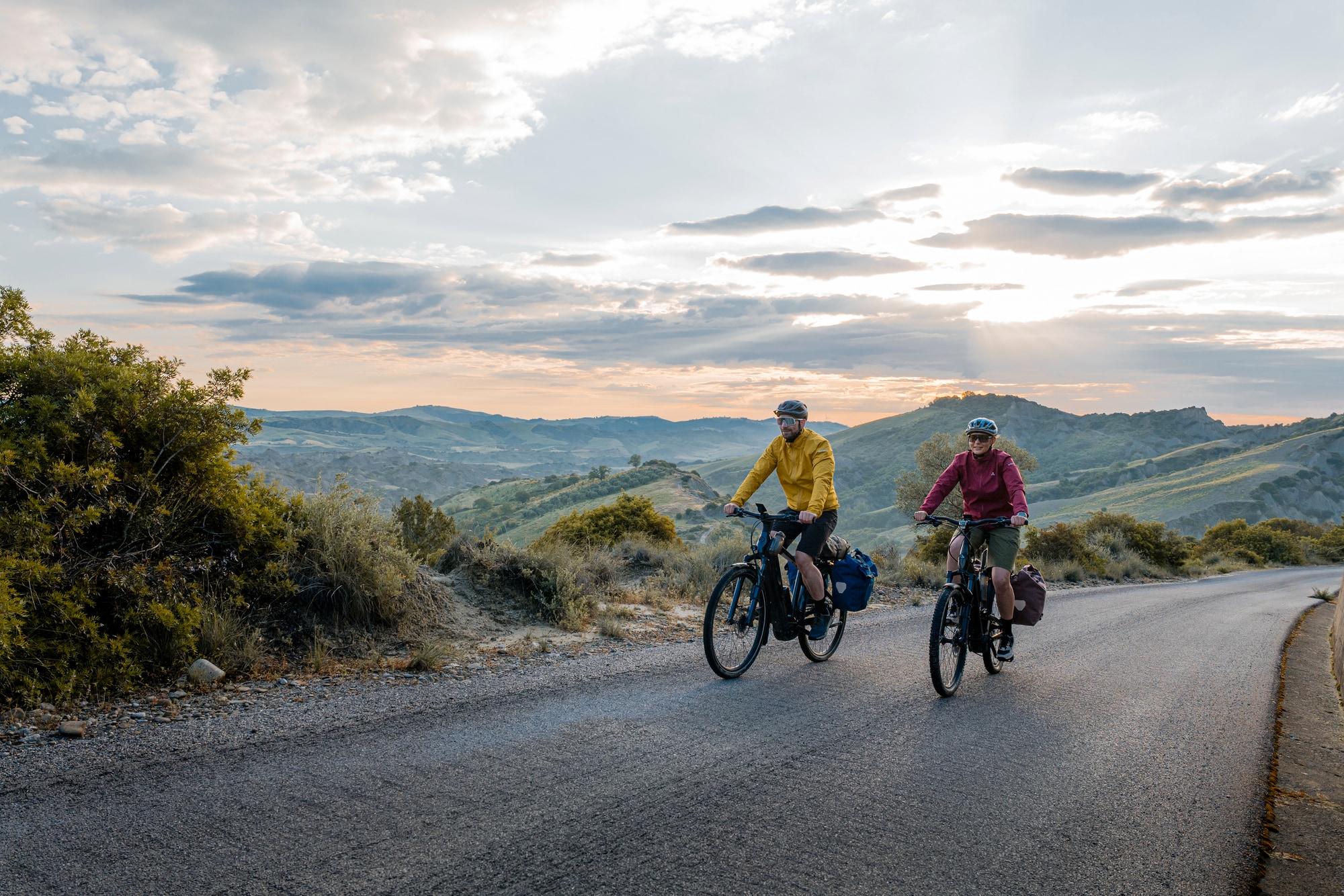 Zwei Personen auf Bosch Performance Line E-Bikes fahren bei Sonnenuntergang durch eine malerische Landschaft.