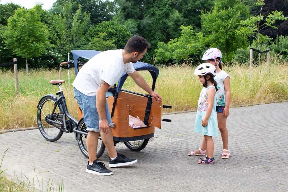 Auf den Bild öffnet ein Mann die Türe der Transportbox für das Babboe Lastenrad, zwei Mädchen stehen davor