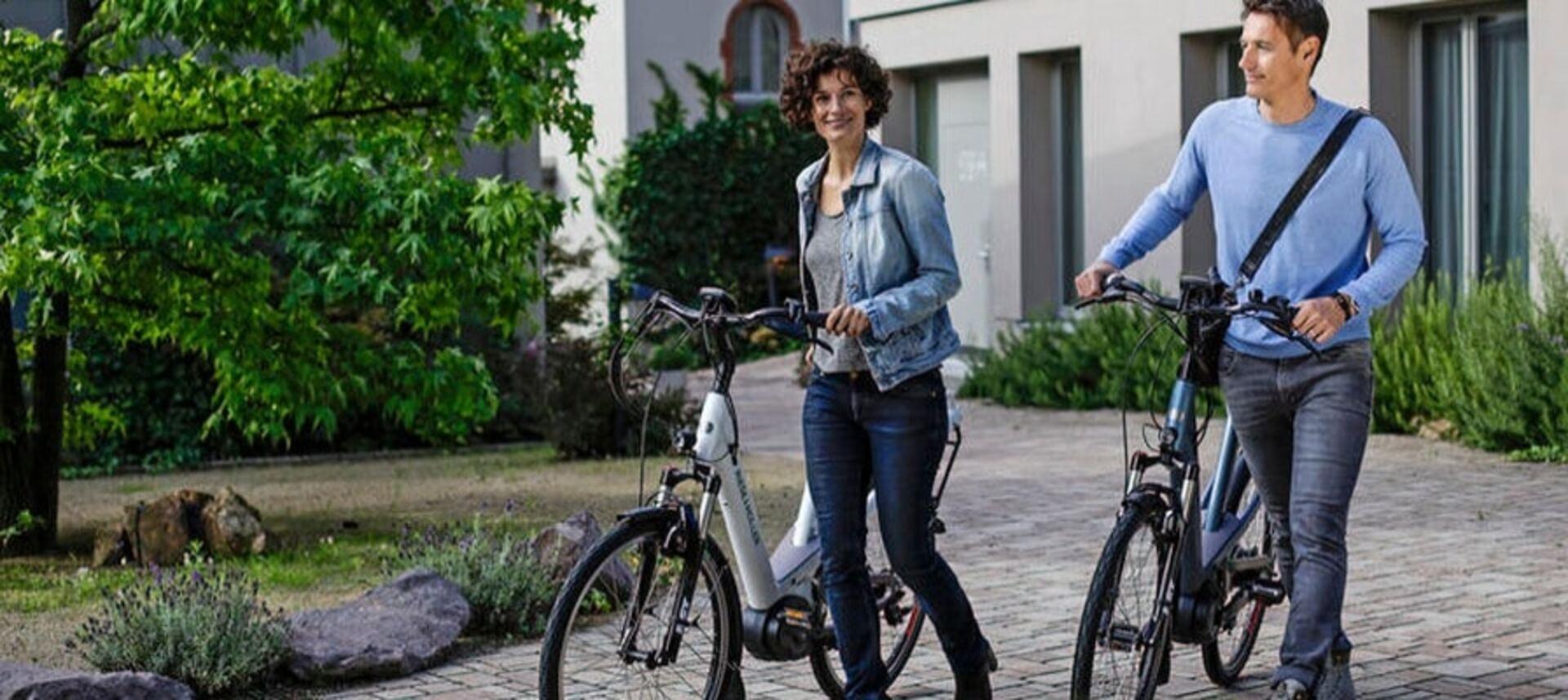 Eine Frau und ein Mann schieben lächelnd ihre Riese & Müller e-Bikes nebeneinander auf einem gepflasterten Weg.