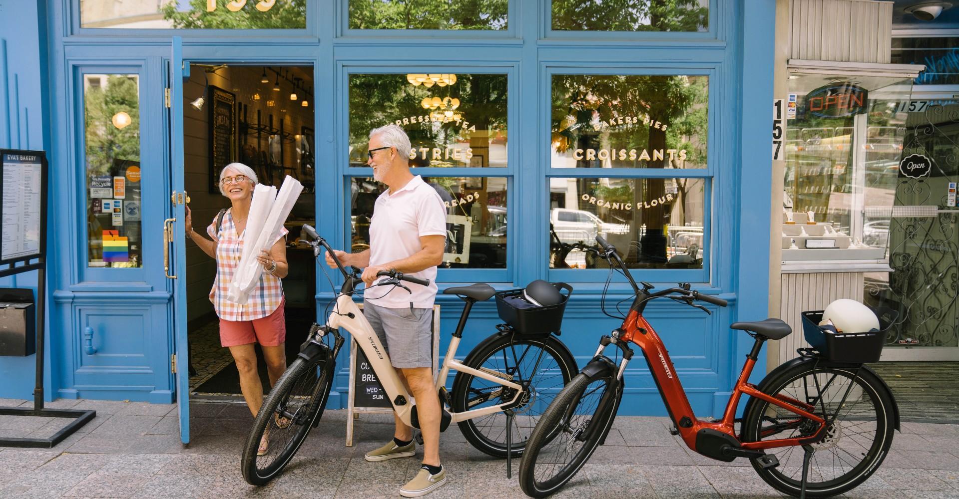 Das Bild zeigt ein Paar älterer Menschen, die vor einem Geschäft stehen und zwei e-Bikes neben sich haben. Beide Fa