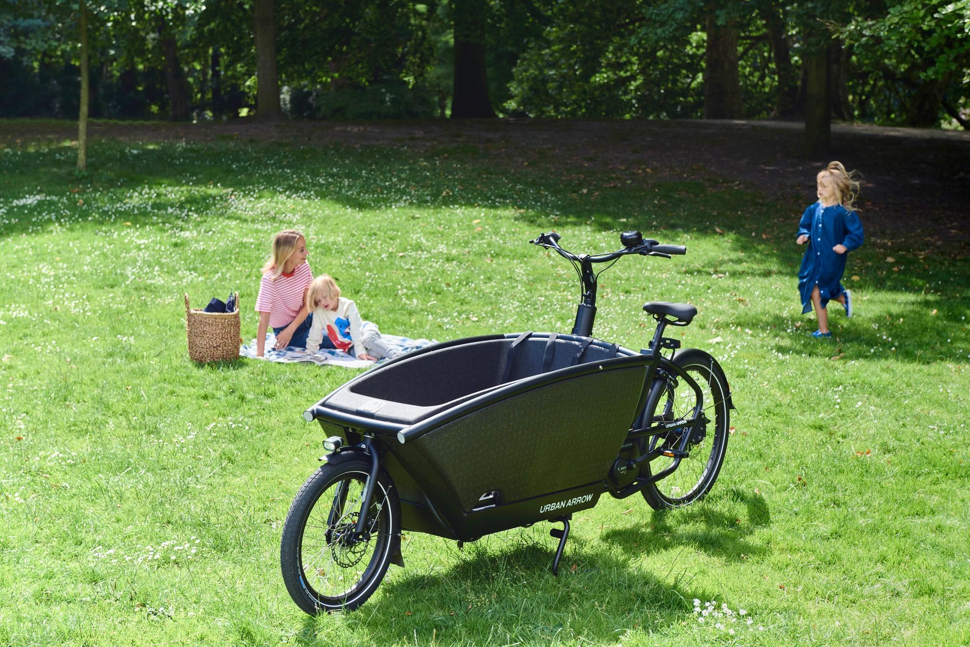 Ein Urban Arrow Family, daneben sitzt eine Mutter mit ihren beiden spielenden Kindern