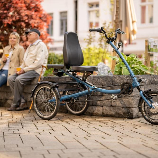 Ein blaues Pfautec-Dreirad steht im Vordergrund, zwei Personen entspannen sich im Hintergrund. Ideal für Mobilität