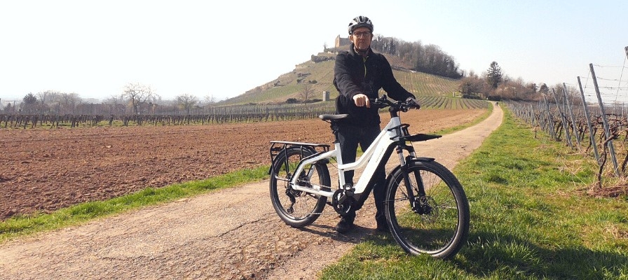 Das Bild zeigt einen Fahrer auf einem modernen E-Bike, das auf einem Schotterweg inmitten von Weinbergen steht.