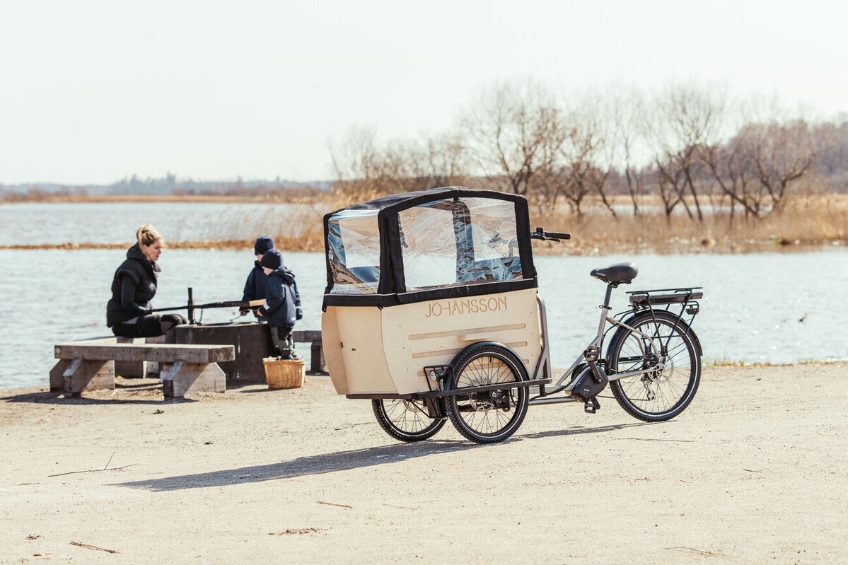 Das Bild zeigt ein E-Bike Lastenrad der Marke Johansson, das am Ufer eines Gewässers abgestellt ist.