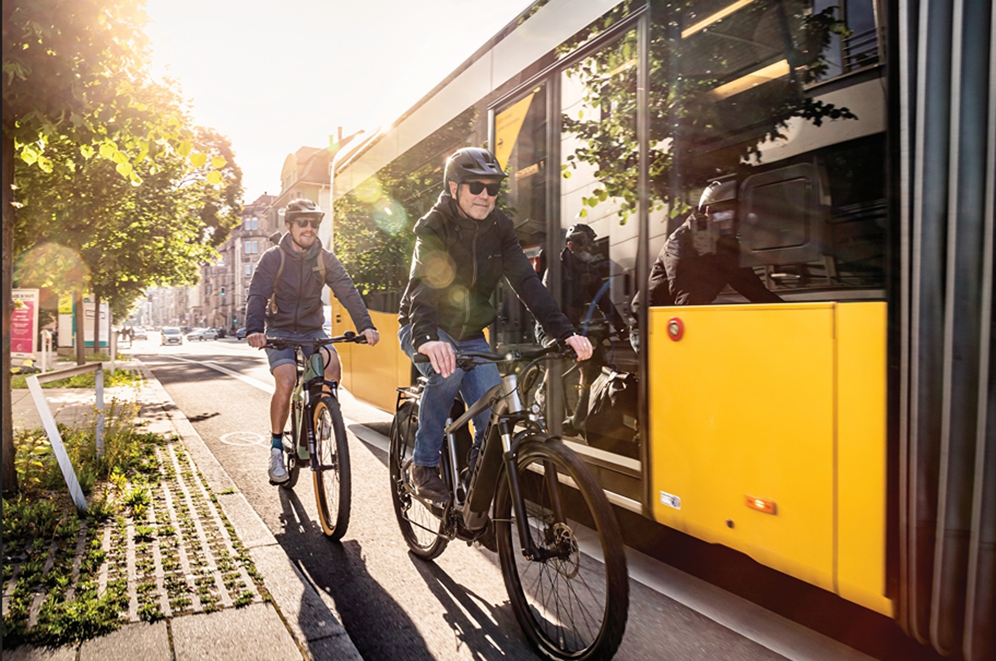 Das Bild zeigt zwei Radfahrer, die auf E-Bikes durch eine Stadt fahren, während ein gelber Bus daneben hält.