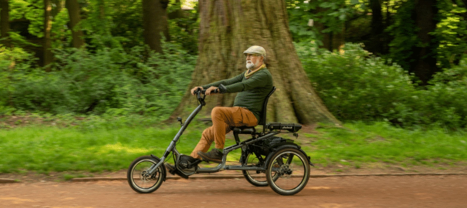 Das Bild zeigt einen älteren Mann, der auf einem pfautec Dreirad sitzt und durch eine parkähnliche Umgebung fährt.