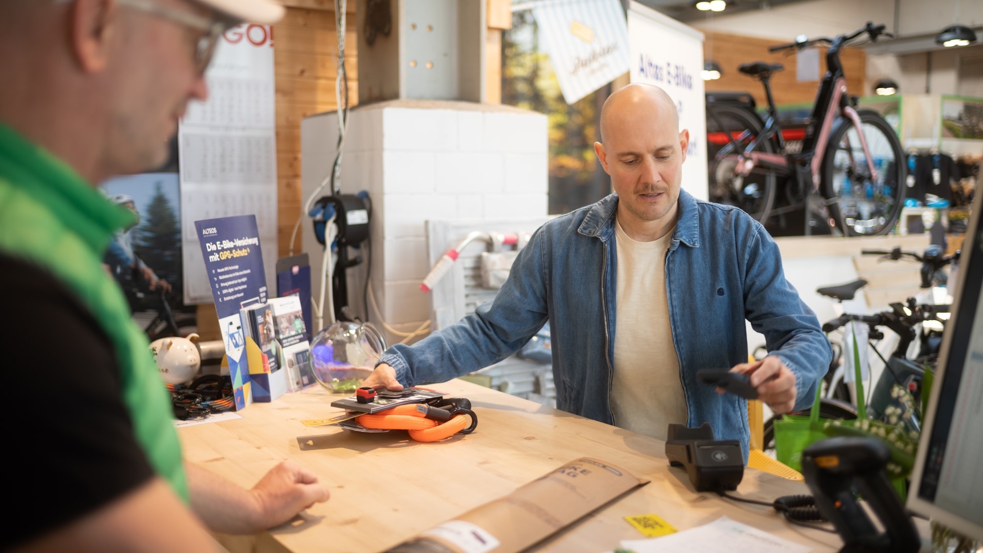 Ein Kunde zahlt Zubehör an der e-motion Kasse in einem E-Bike-Geschäft. Verschiedene E-Bikes und Zubehör sind im Hi