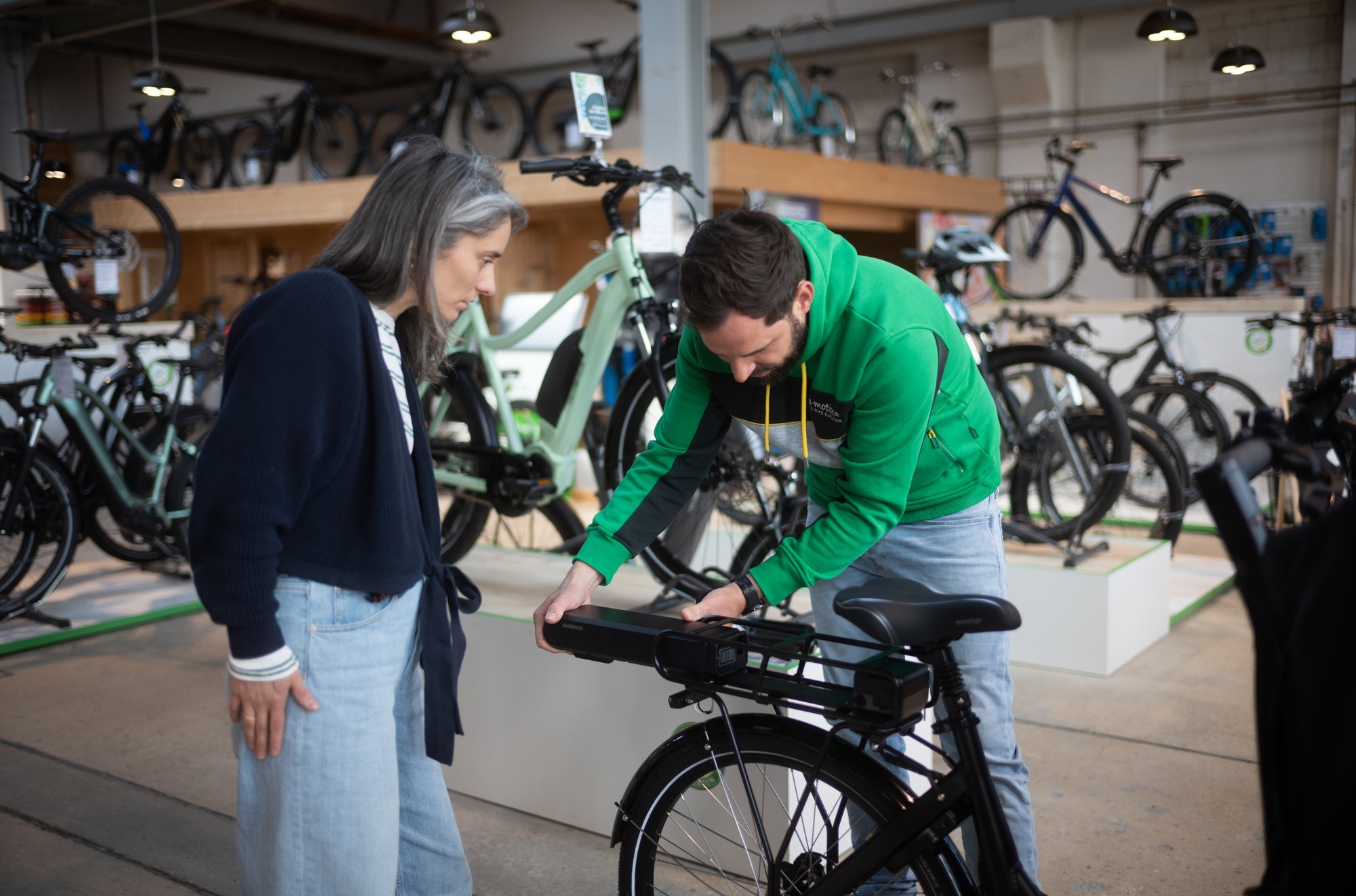 Eine Frau und ein Mann interagieren mit einem e-Bike in einem Fahrradladen. Der Mann in einem grünen Hoodie zeigt d