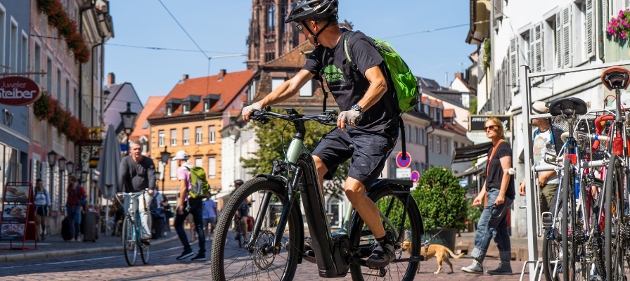 Ein Mann auf einem E-Bike fährt durch eine belebte Stadtstraße mit historischer Architektur im Hintergrund.