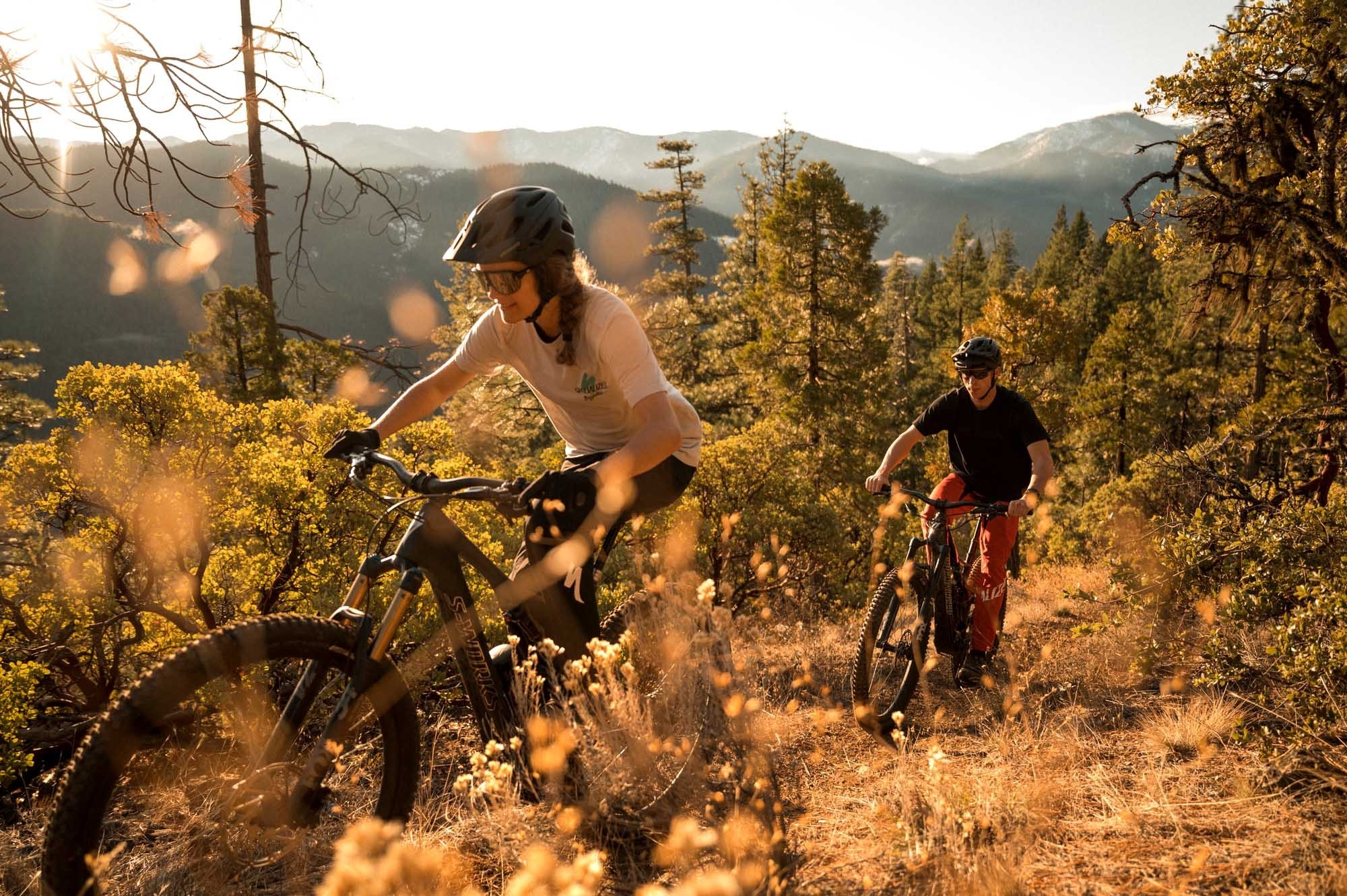 Das Bild zeigt zwei Radfahrer, die auf einem Trail in einer bewaldeten, bergigen Umgebung fahren.