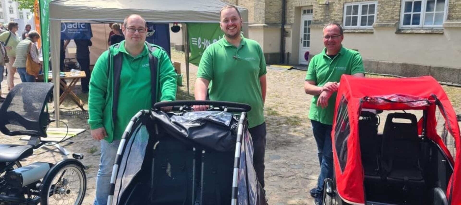 Das Bild zeigt drei Männer in grünen T-Shirts, die am Freiluft-Ökomarkt stehen.